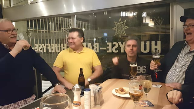 Lachende Stammgäste am Tisch in der Huefyse-Bar, im Fenster der Schriftzug Huefyse-Bar